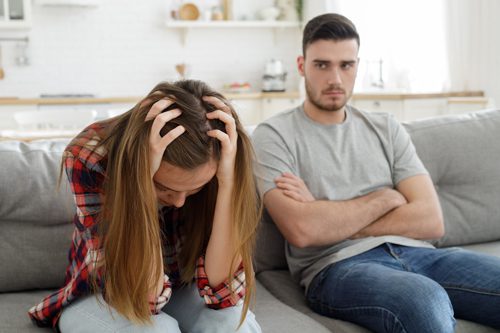woman with hands in her hair, frustrated, next to upset man on couch - spouse's addiction