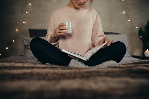girl sitting cross-legged on her bed drinking coffee and reading a book - routines