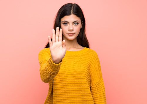 beautiful young woman in golden colored sweater holding her arm out in front of her in a stop motion on a peach background - support