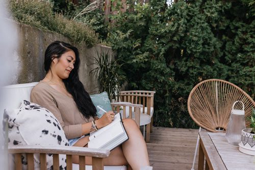 beautiful young woman with long dark hair sitting outside in her backyard writing in a journal - mindfulness